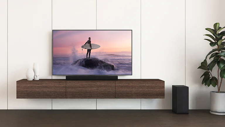Телевизор и саундбар LG размещены на коричневой полке, а сабвуфер — на полу. На экране телевизора изображен серфер на скале.