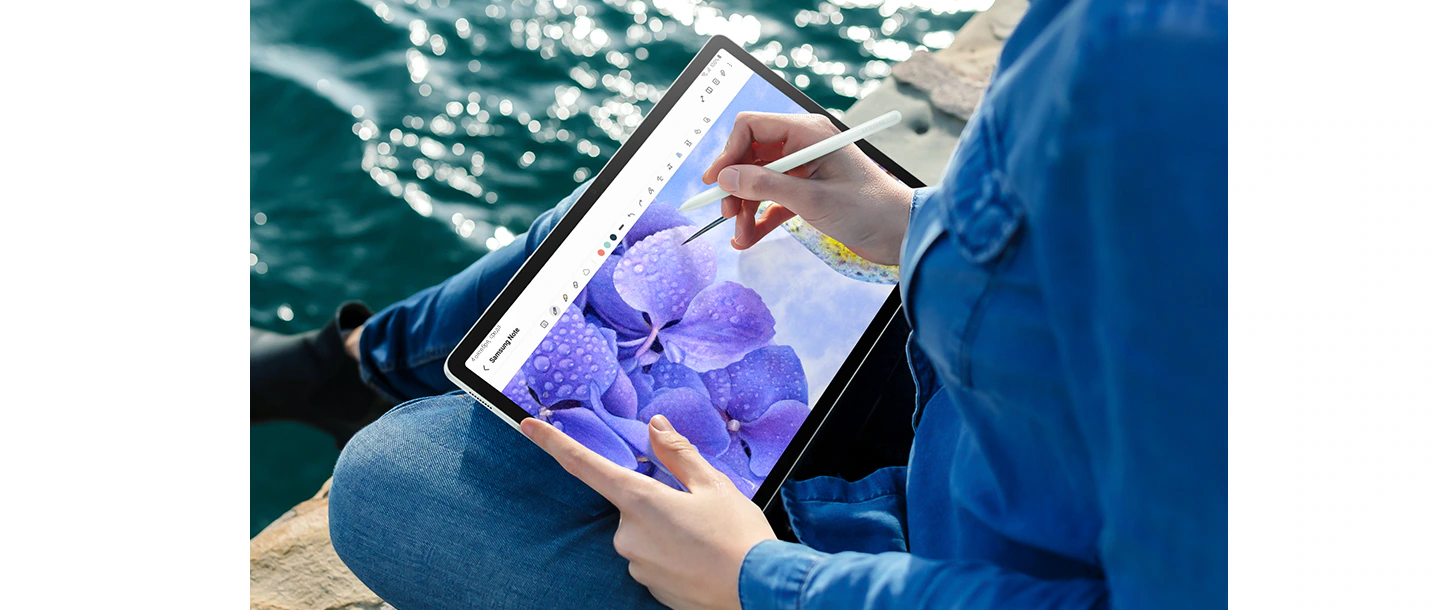 Человек, сидящий у воды, держит в руках Galaxy Tab S9 FE+ в горизонтальном режиме и пером S Pen рисует цветок в открытом на экране приложении Samsung Notes.